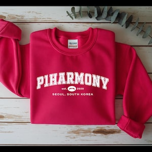 Może przedstawiać: Żywa czerwona bluza z białym napisem "PIHARMONY" w łukowatym kształcie. Poniżej widnieje napis "est. 2020" i "SEOUL, SOUTH KOREA". Bluza jest ułożona na jasnej powierzchni imitującej drewno.