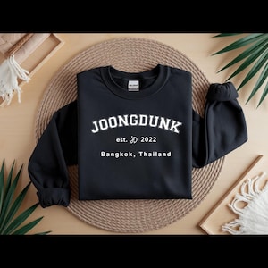 May include: Black crewneck sweatshirt with the word "JOONGDUNK" in white block letters. Below the name is "est. 2022" and "Bangkok, Thailand" in a smaller font. The sweatshirt is displayed on a woven mat.