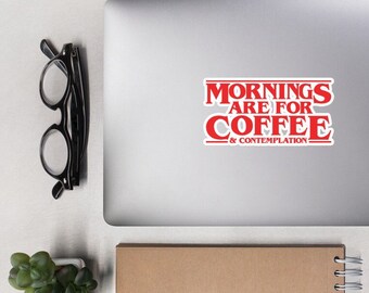 May include: A silver laptop with a red sticker that says "Mornings are for Coffee & Contemplation". The laptop is on a white surface with a brown notebook, a white mouse, and two small potted plants.