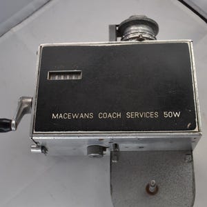 May include: A vintage mechanical ticket machine with a black rectangular face featuring the white text MACEWANS COACH SERVICES 50W. The device includes a silver metal body, a side-mounted hand crank with a black handle, and a numerical counter display.