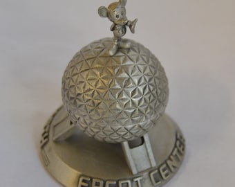 Könnte beinhalten: Eine silberfarbene Epcot Center Souvenirfigur. Die Figur zeigt eine geometrische Kugel mit einer kleinen Mickey Mouse Figur oben. Auf dem Sockel der Figur steht "EPCOT CENTER".