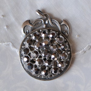 May include: A silver pendant featuring a round design of black rhinestones. The pendant has a decorative, ornate bail.