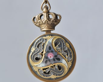 May include: A gold pendant with a crown and a round, gray, and gold enamel design. The design features a stylized floral pattern and a pink and white center.