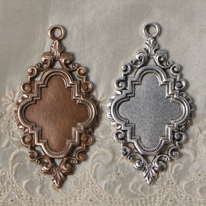 May include: Two ornate metal pendants, one silver and one bronze, with a four-leaf clover design. The pendants are on a white lace background.