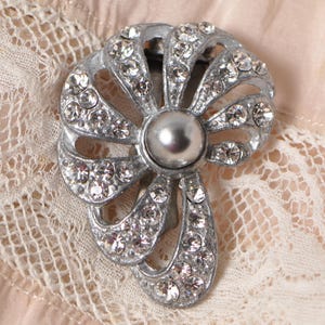 May include: Vintage silver brooch featuring a central faux pearl and sparkling clear rhinestones. The brooch has a fan-shaped design with openwork details. The rhinestones are set in silver metal, creating a shimmering effect. A classic piece of jewellery.