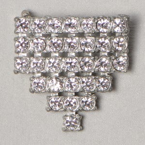 May include: A silver brooch with a geometric design. The brooch features multiple rows of clear, round rhinestones, arranged in a rectangular shape with a descending, triangular bottom. The pin is set against a light grey background.