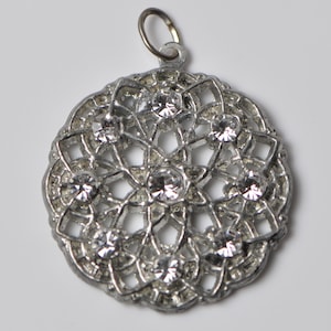 May include: Silver-toned pendant with a circular, openwork design. The pendant features a series of clear, round gemstones set within the intricate metalwork. A small loop is at the top for attaching to a chain. The pendant is likely a piece of jewelry.