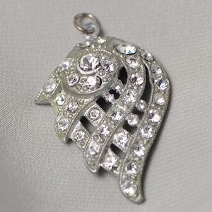 Puede incluir: Colgante plateado con diseño de ala, incrustado con numerosas piedras de estrás claras y brillantes. Tiene un pequeño lazo en la parte superior para una cadena. El fondo es de tela gris claro.