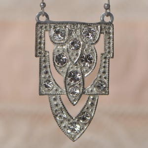 May include: Silver-toned pendant necklace with a geometric design. The pendant features a series of clear, round rhinestones set within an Art Deco-inspired frame. The necklace hangs from a delicate silver chain, creating a vintage-inspired accessory.