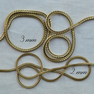 May include: Two gold braided cords, one 2 mm and one 3 mm in diameter, are shown on a white fabric background. The cords are arranged in a decorative pattern.