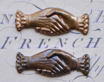 Puede incluir: Dos apretones de manos de metal bronce antiguo, cada uno con un diseño detallado de manos entrelazadas. Las manos están adornadas con detalles decorativos y tienen una estética vintage.