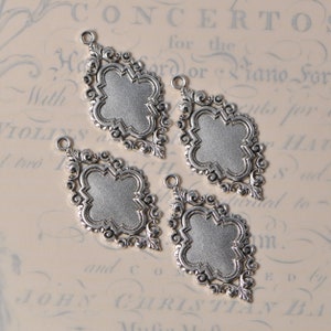 May include: Four silver-toned metal charms with a decorative filigree design and a blank space in the center. The charms are shaped like a diamond with rounded corners.