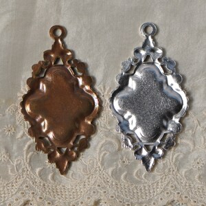 May include: Two ornate metal pendants, one gold and one silver, with a decorative, intricate design. Both pendants have a blank center space for adding a design or image.