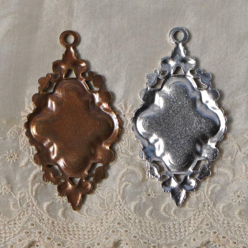 May include: Two ornate metal pendants, one gold and one silver, with a decorative, intricate design. Both pendants have a blank center space for adding a design or image.
