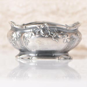 Puede incluir: Un cuenco plateado con borde festoneado y detalles florales en relieve. El cuenco presenta una base de pedestal y est&aacute; adornado con intrincados patrones de hojas y flores. La superficie reflectante debajo refleja el dise&ntilde;o del cuenco.