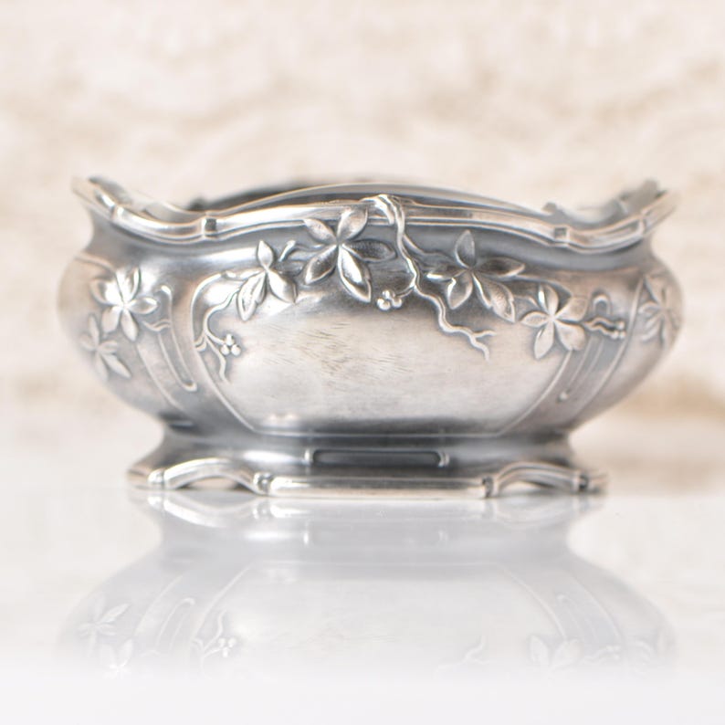 Puede incluir: Un cuenco plateado con borde festoneado y detalles florales en relieve. El cuenco presenta una base de pedestal y est&aacute; adornado con intrincados patrones de hojas y flores. La superficie reflectante debajo refleja el dise&ntilde;o del cuenco.
