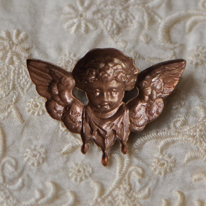May include: A copper-coloured angel figurine with wings and a cherubic face. The angel is depicted wearing a flowing robe and has a delicate, vintage aesthetic.