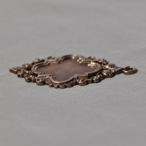 May include: A decorative metal pendant with an ornate design. The pendant has a four-leaf clover shape with a central oval space. The pendant is made of a dark brown metal with a shiny finish.