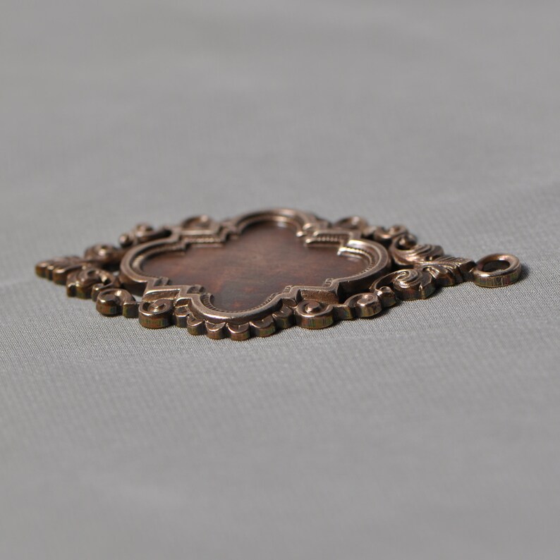 May include: A decorative metal pendant with an ornate design. The pendant has a four-leaf clover shape with a central oval space. The pendant is made of a dark brown metal with a shiny finish.