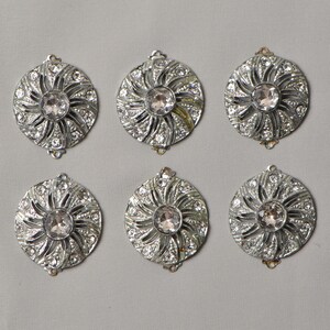 May include: Six silver-toned, circular embellishments with a central clear rhinestone surrounded by smaller clear rhinestones. The design features a sunburst pattern. Each piece has a small loop at the top for attachment.