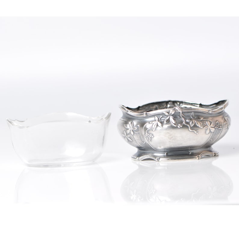 Puede incluir: Dos cuencos decorativos. Uno es de vidrio transparente, el otro de metal plateado. El cuenco de metal tiene un dise&ntilde;o floral y de vid. Ambos cuencos tienen un borde festoneado. Los cuencos est&aacute;n sobre una superficie blanca.