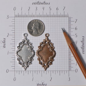 May include: Two ornate metal pendants, one silver and one copper, with a quatrefoil design. The pendants are approximately 2 inches in length and are shown next to a ruler and a US quarter for scale.