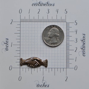 May include: A bronze-colored metal handshake charm, approximately 1 inch in length, with a textured surface. The charm is shown next to a US quarter for size comparison.