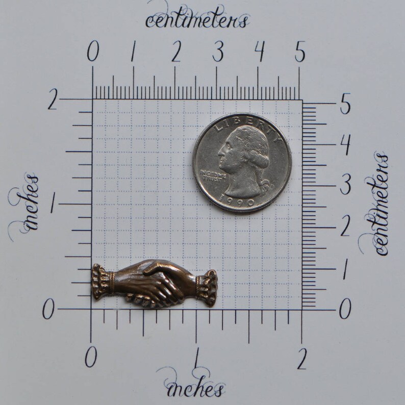 May include: A bronze-colored metal handshake charm, approximately 1 inch in length, with a textured surface. The charm is shown next to a US quarter for size comparison.