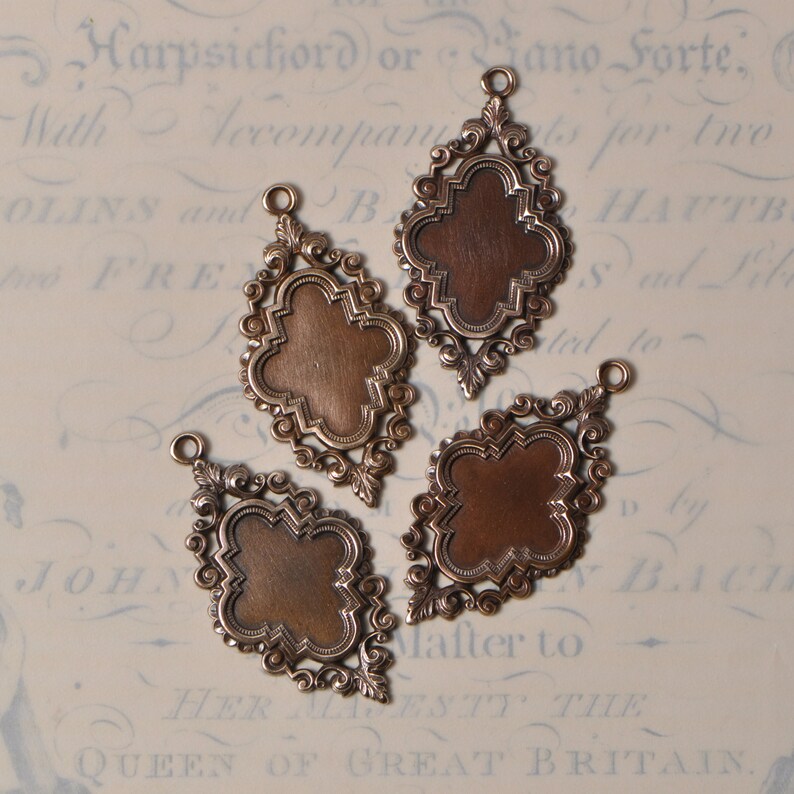 May include: Three antique-style metal charms with ornate filigree designs. Each charm features a blank, copper-colored oval in the center.