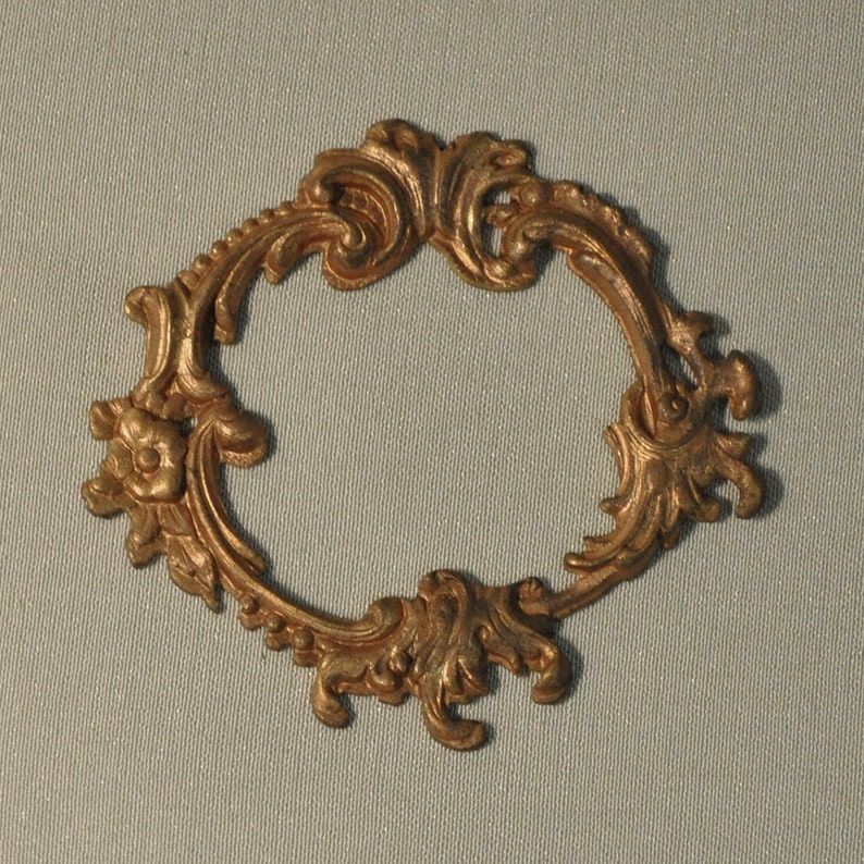 May include: A gold-colored metal frame with a decorative, ornate design. The frame is oval-shaped and has a smooth, polished finish.