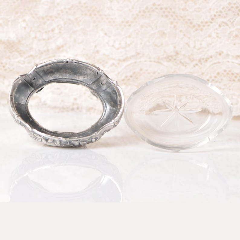 Puede incluir: Un collar y un tap&oacute;n de botella de perfume plateados antiguos. El collar tiene un dise&ntilde;o floral alrededor del borde. El tap&oacute;n de vidrio transparente tiene un dise&ntilde;o de estrella. Los art&iacute;culos est&aacute;n sobre una superficie reflectante.