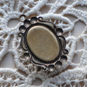May include: A bronze-colored metal frame with an oval opening, perfect for adding a cameo or other decorative element. The frame has a decorative design with a series of small, raised circles around the edge.