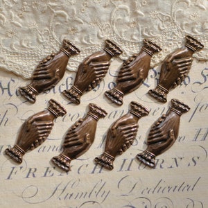 May include: Eight antique bronze metal charms shaped like two hands clasped together. The charms are detailed with a textured surface and have a slightly aged look.