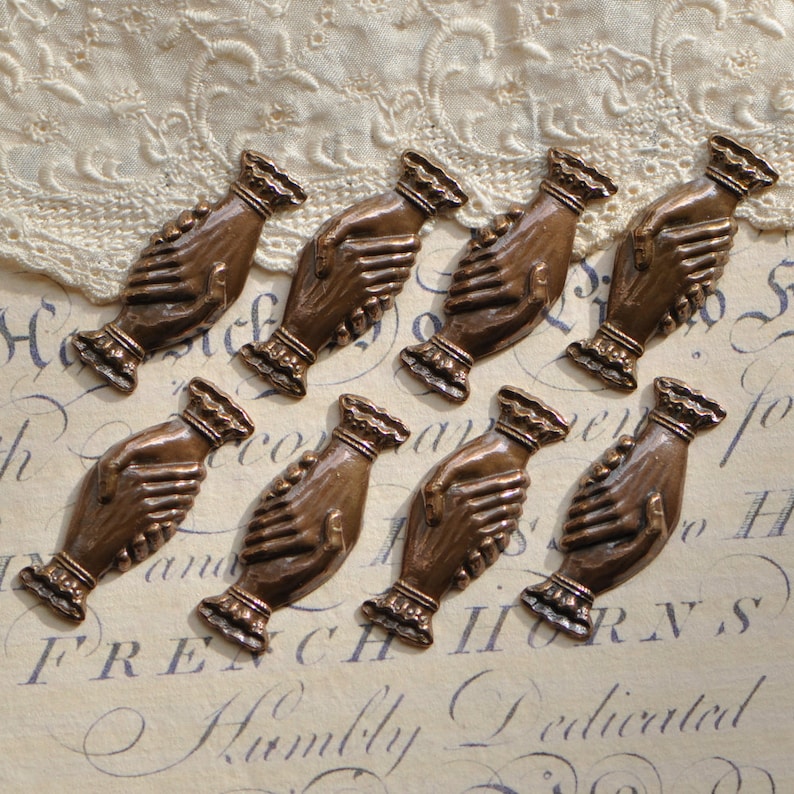 May include: Eight antique bronze metal charms shaped like two hands clasped together. The charms are detailed with a textured surface and have a slightly aged look.
