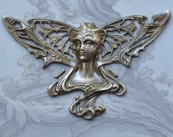 May include: A silver-toned Art Nouveau style decorative piece featuring a woman's face with ornate wings. The design includes intricate details and flowing lines, reminiscent of a butterfly or mythical creature. The piece is set against a light blue and gray background.