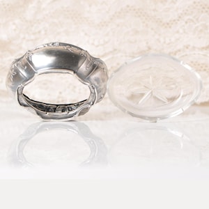 Puede incluir: Un servilletero plateado con detalles florales y un plato de vidrio transparente con un dise&ntilde;o de estrella. Los objetos se colocan sobre un fondo blanco roto suave, creando una est&eacute;tica vintage.