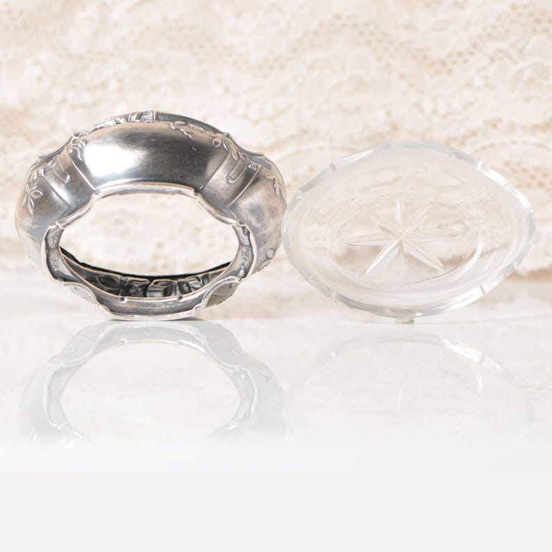 Puede incluir: Un servilletero plateado con detalles florales y un plato de vidrio transparente con un dise&ntilde;o de estrella. Los objetos se colocan sobre un fondo blanco roto suave, creando una est&eacute;tica vintage.