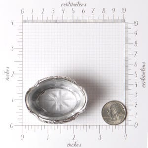 Puede incluir: Un peque&ntilde;o plato ovalado de plata con un borde decorativo. El plato mide aproximadamente 7,6 cm de largo y 5,1 cm de ancho. Una moneda est&aacute; al lado del plato para comparar el tama&ntilde;o. El plato tiene un dise&ntilde;o de estrella en el centro.