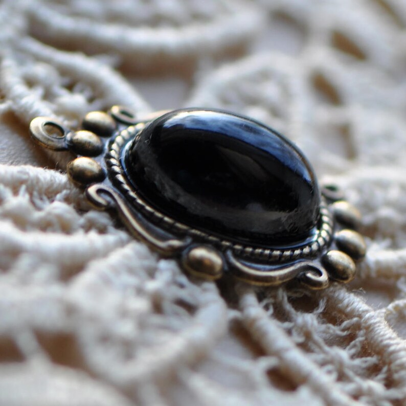 May include: A black oval-shaped gemstone set in a silver-toned ornate frame with decorative details. The frame has a beaded edge and a textured surface.