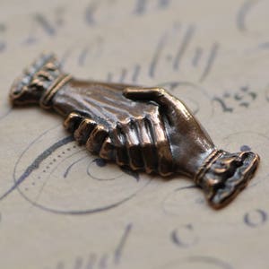 May include: A bronze sculpture of two hands clasped together in a handshake. The sculpture is detailed and realistic, with visible wrinkles and lines on the hands.