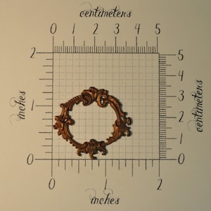 May include: A gold-colored metal frame with an ornate design. The frame is approximately 2 inches in diameter.