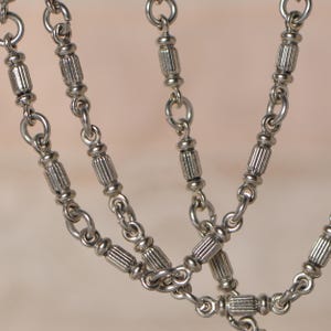 Könnte beinhalten: Nahaufnahme einer silberfarbenen Halskette mit mehreren Strängen. Die Halskette hat kleine, dekorative zylindrische Perlen und verbindende Kettenglieder. Das Design hat eine Vintage- oder Antik-Ästhetik.