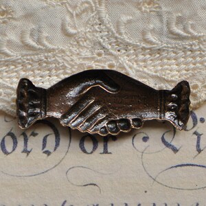 May include: A bronze-colored metal pin shaped like two hands shaking. The pin is detailed with textured lines and a raised design.