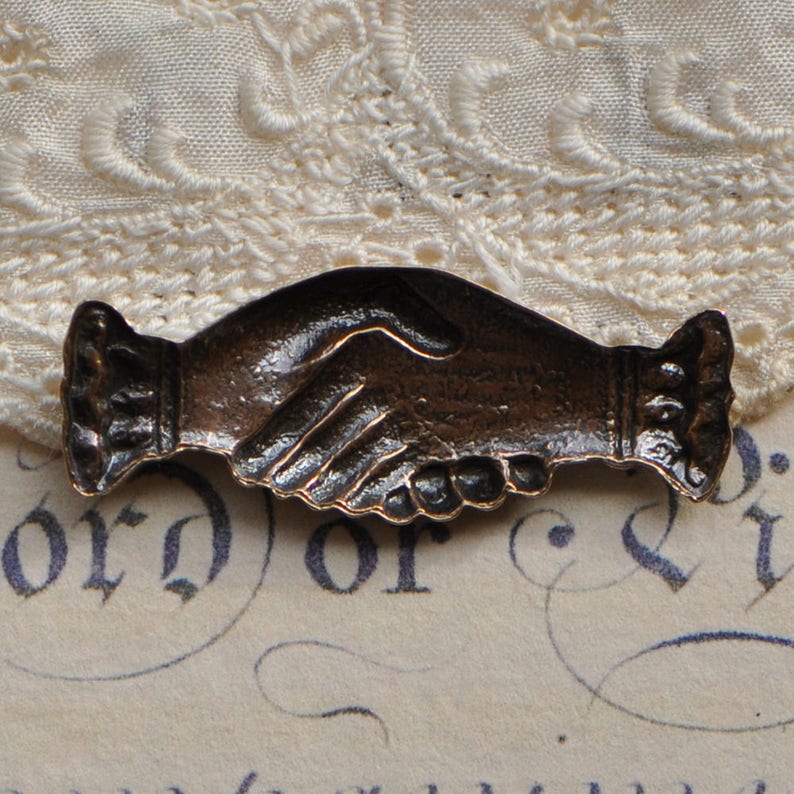 May include: A bronze-colored metal pin shaped like two hands shaking. The pin is detailed with textured lines and a raised design.