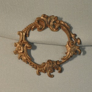 May include: A gold-toned metal frame with an ornate, scrolled design. The frame is oval-shaped and has a delicate, intricate pattern.