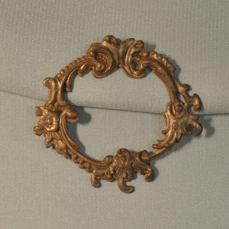 May include: A gold-toned metal frame with an ornate, scrolled design. The frame is oval-shaped and has a delicate, intricate pattern.