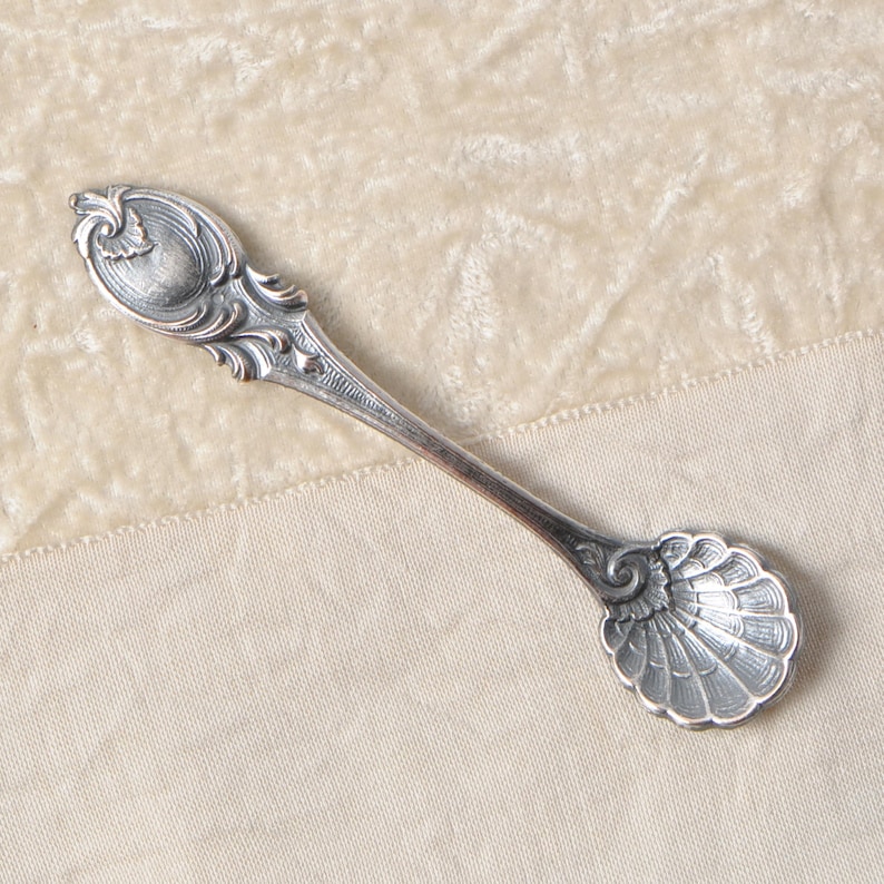 Puede incluir: Una cuchara de plata con detalles ornamentados. La cuchara presenta un cuenco en forma de concha y un mango con un dise&ntilde;o ovalado. La cuchara es probablemente vintage o antigua, con un acabado deslustrado. La cuchara est&aacute; sobre una superficie color crema.