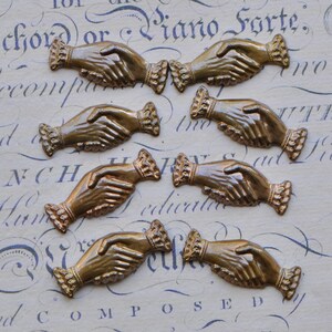 May include: Seven antique brass hand-shaped charms with a detailed design. Each charm features two hands clasped together, symbolizing friendship or unity.