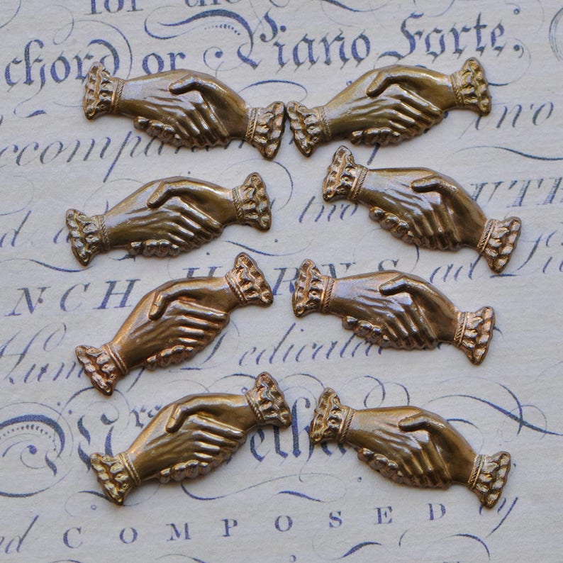 May include: Seven antique brass hand-shaped charms with a detailed design. Each charm features two hands clasped together, symbolizing friendship or unity.