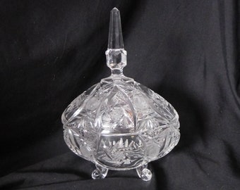 May include: A clear glass candy dish with a domed lid and three legs. The dish is decorated with a cut glass pattern.